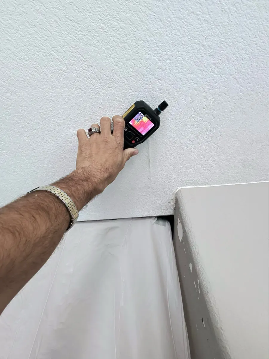 Thermal moisture meter held against a white wall above plastic sheeting in The Lakes home